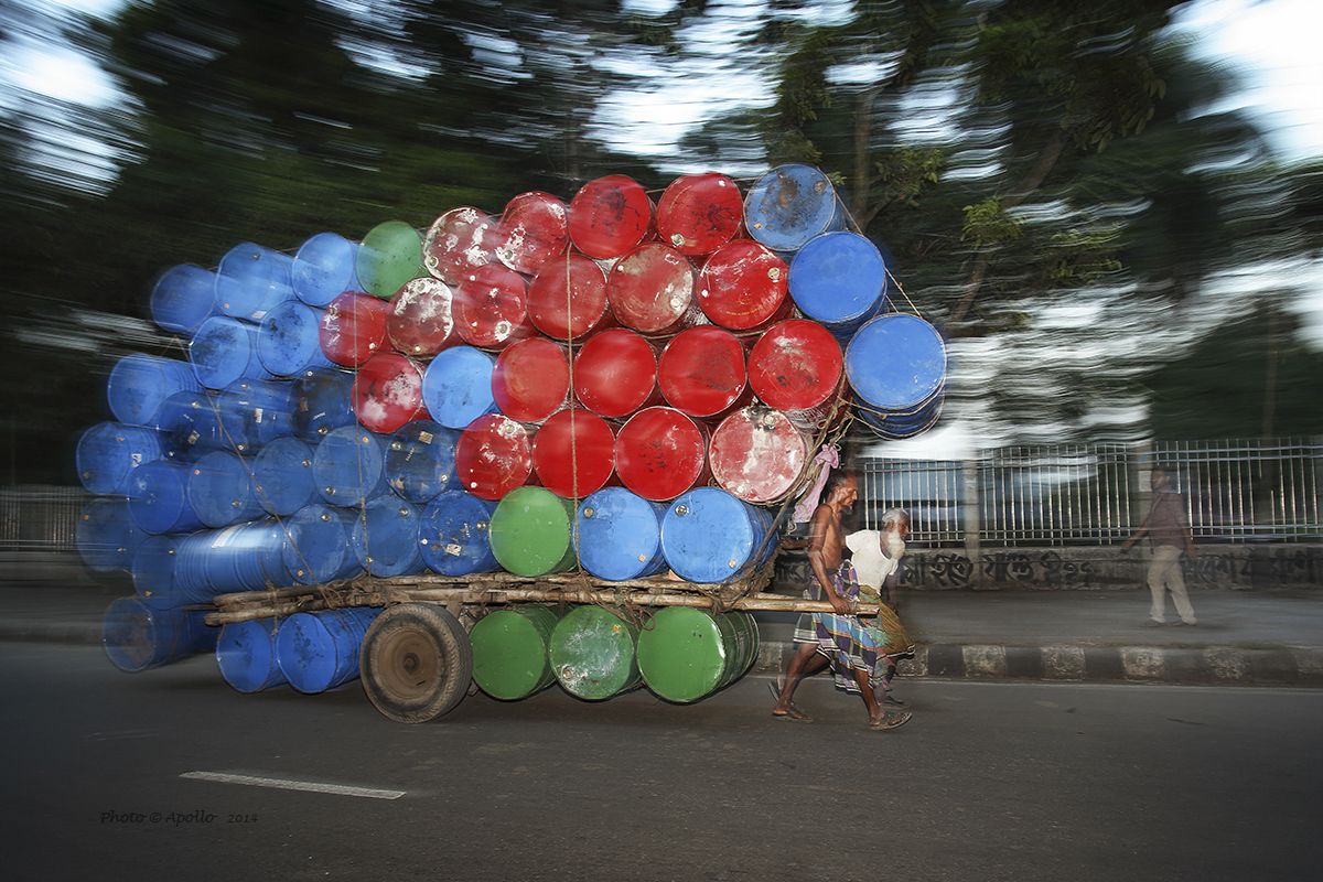 Two men are carrying over loaded empty metal drums on a push cart. 